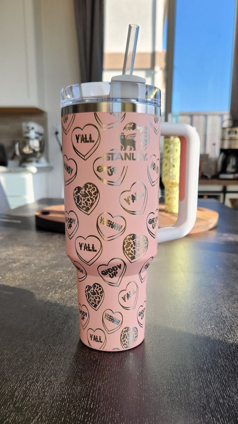 May include: Pink stainless steel tumbler with a white lid and a straw. The tumbler has a gold print of hearts with various sayings, including "Y'all", "Giddy Up", and "Yeehaw". The tumbler also has the Stanley logo printed on it.