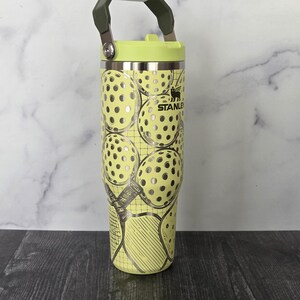 May include: A stainless steel Stanley tumbler with a light green lid and handle. The tumbler is decorated with a pattern of pickleball graphics in silver and yellow. The Stanley logo is visible.