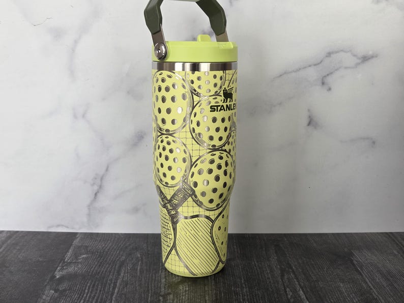 May include: A stainless steel Stanley tumbler with a light green lid and handle. The tumbler is decorated with a pattern of pickleball graphics in silver and yellow. The Stanley logo is visible.