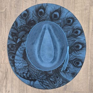 May include: A blue fedora hat with a peacock design. The hat features a detailed illustration of a peacock and its feathers in black against a blue background. A blue tassel hangs from the brim.