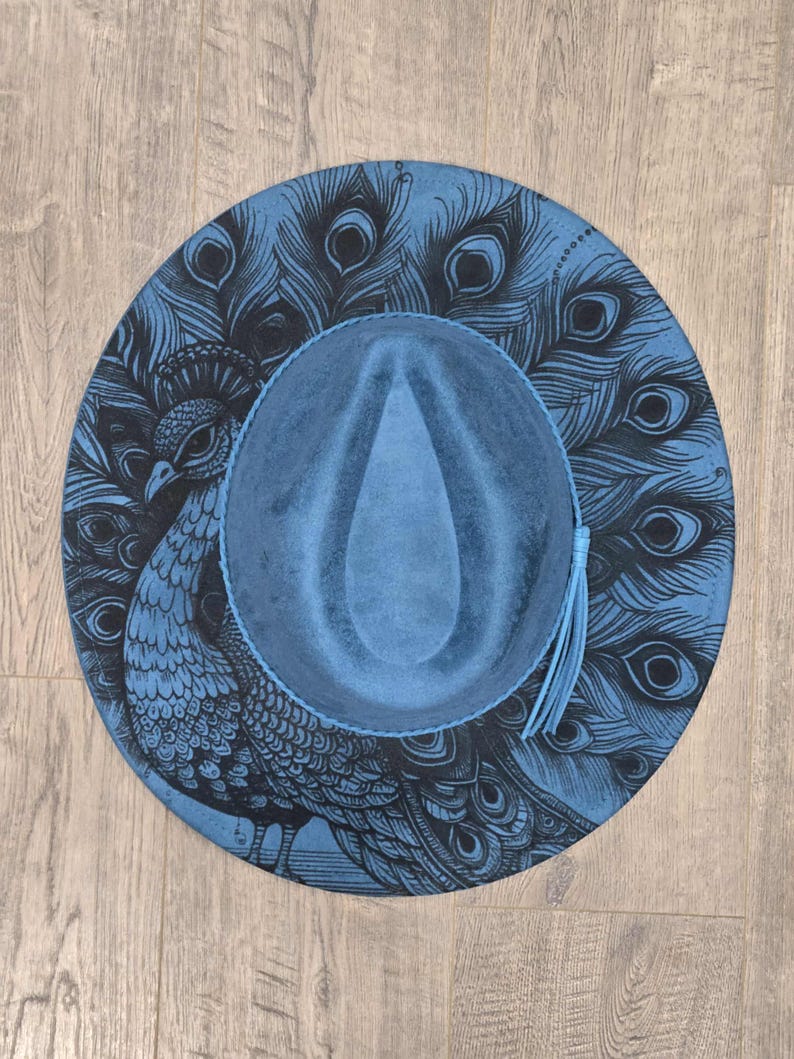 May include: A blue fedora hat with a peacock design. The hat features a detailed illustration of a peacock and its feathers in black against a blue background. A blue tassel hangs from the brim.