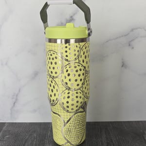 May include: A yellow tumbler with a lime green lid and handle. The tumbler is decorated with a pattern of pickleballs and a pickleball racket. The handle is white and gray.