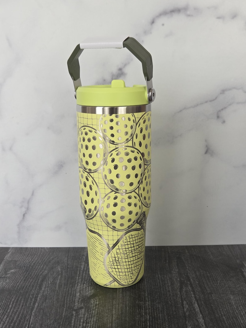 May include: A yellow tumbler with a lime green lid and handle. The tumbler is decorated with a pattern of pickleballs and a pickleball racket. The handle is white and gray.