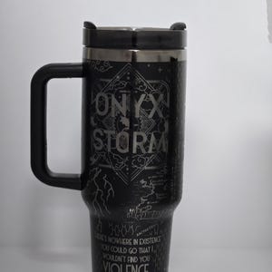 May include: Black travel mug with a black handle and lid. The mug features the text "ONYX STORM" and other text and graphics. The bottom of the mug has the text "THERE'S NOWHERE IN EXISTENCE YOU COULD GO THAT WOULDN'T FIND YOU VIOLENCE ARETIA."