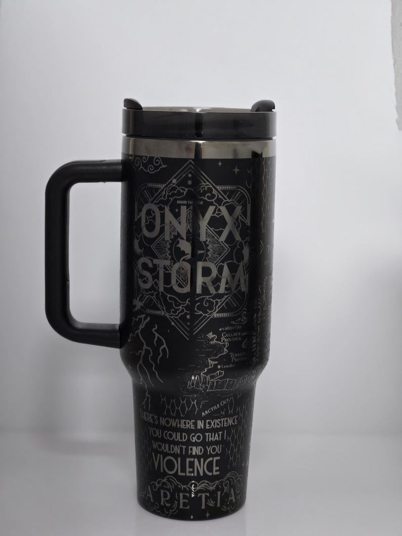 May include: Black travel mug with a black handle and lid. The mug features the text "ONYX STORM" and other text and graphics. The bottom of the mug has the text "THERE'S NOWHERE IN EXISTENCE YOU COULD GO THAT WOULDN'T FIND YOU VIOLENCE ARETIA."