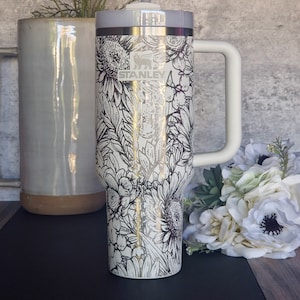 Könnte beinhalten: Eine weiße Edelstahl-Reisebecher mit einem schwarzen Blumendesign und einem schwarzen Griff. Der Becher hat einen silbernen Deckel und das Wort "STANLEY" ist auf der Seite aufgedruckt.