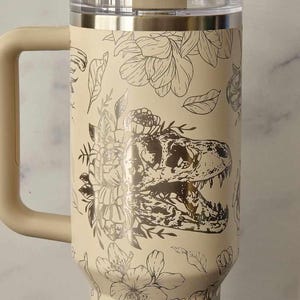 Puede incluir: Una taza de viaje de acero inoxidable de color crema con un dise&ntilde;o floral y de dinosaurio en blanco y negro. La taza tiene un asa y una tapa.