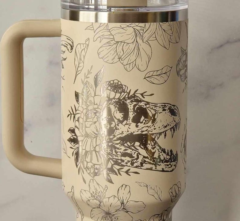 Puede incluir: Una taza de viaje de acero inoxidable de color crema con un dise&ntilde;o floral y de dinosaurio en blanco y negro. La taza tiene un asa y una tapa.