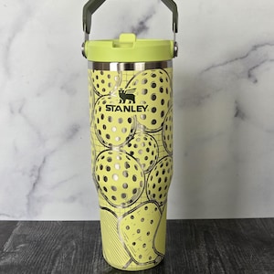 May include: A yellow Stanley tumbler with a pickleball design. The tumbler has a green handle and lid. The pickleball design is silver and black. The tumbler has the Stanley logo.