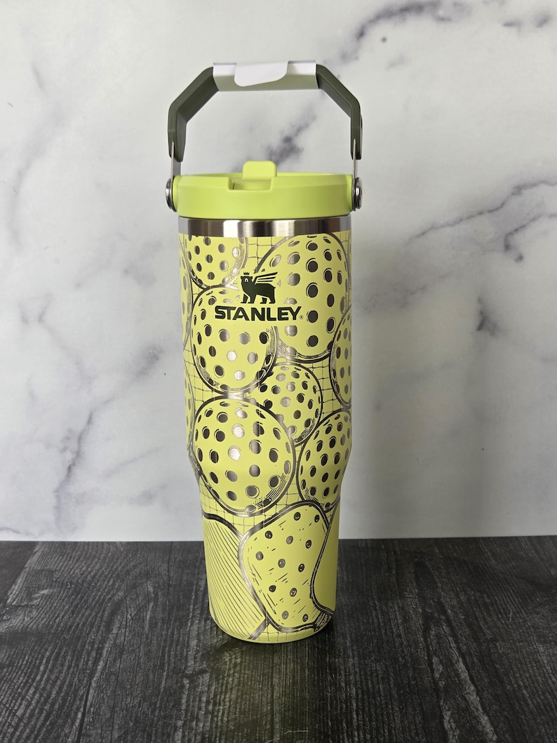 May include: A yellow Stanley tumbler with a pickleball design. The tumbler has a green handle and lid. The pickleball design is silver and black. The tumbler has the Stanley logo.