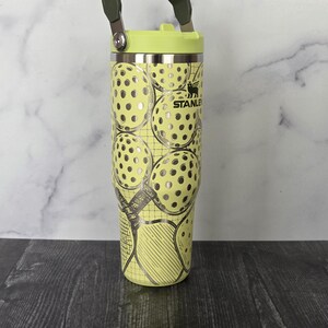 May include: A yellow Stanley tumbler with a green handle and lid. The tumbler features a pattern of tennis balls with silver accents. The Stanley logo is visible on the side.