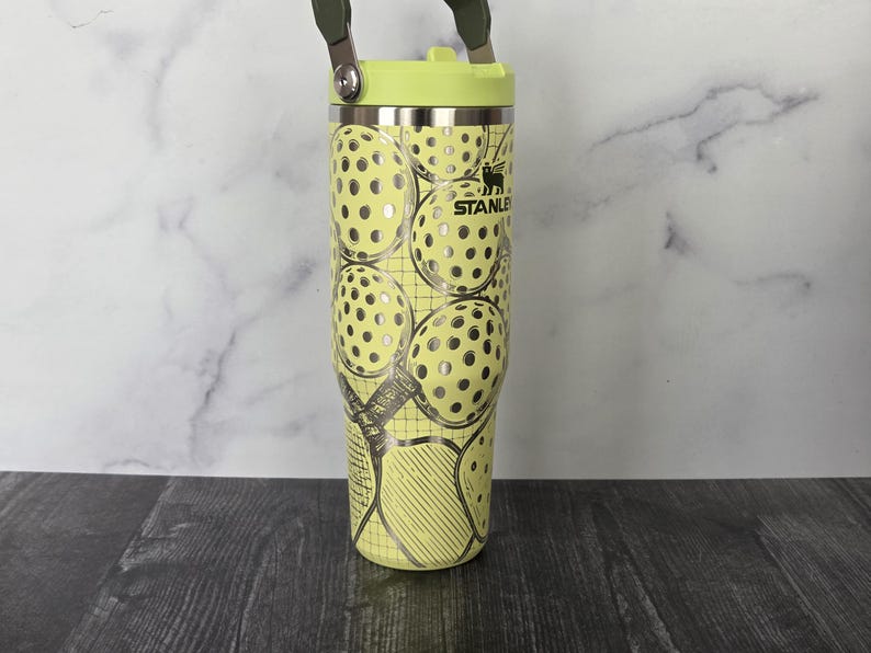 May include: A yellow Stanley tumbler with a green handle and lid. The tumbler features a pattern of tennis balls with silver accents. The Stanley logo is visible on the side.