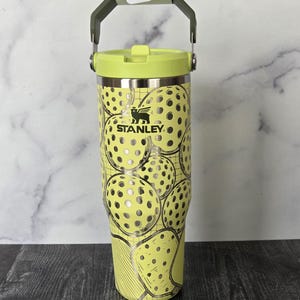 May include: A yellow Stanley tumbler with a lime green lid and handle. The tumbler is decorated with a pattern of pickleball graphics. The Stanley logo is visible on the front of the tumbler. The tumbler has a capacity of approximately 30 ounces.