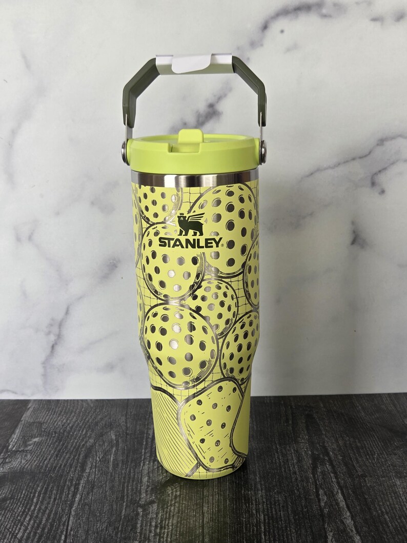 May include: A yellow Stanley tumbler with a lime green lid and handle. The tumbler is decorated with a pattern of pickleball graphics. The Stanley logo is visible on the front of the tumbler. The tumbler has a capacity of approximately 30 ounces.