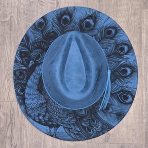 May include: A blue fedora hat with a peacock feather design. The hat has a blue crown and brim, with a black peacock illustration on the brim. A blue tassel hangs from the side. The hat is a stylish accessory.