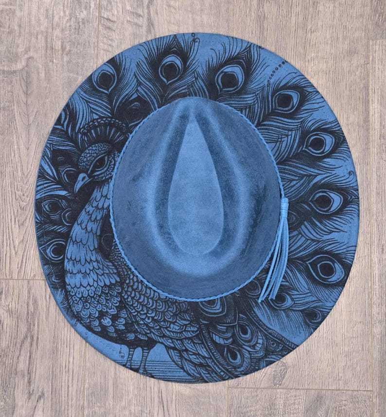 May include: A blue fedora hat with a peacock feather design. The hat has a blue crown and brim, with a black peacock illustration on the brim. A blue tassel hangs from the side. The hat is a stylish accessory.