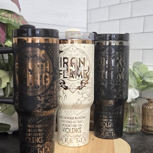 May include: Three travel mugs with black, white, and gold designs. The black and gold mugs have intricate patterns and text. The white mug features a map and the words "Iron Flame." All mugs have black lids and are displayed on a wooden tray.