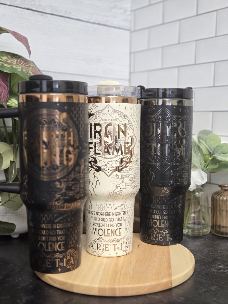 May include: Three travel mugs with black, white, and gold designs. The black and gold mugs have intricate patterns and text. The white mug features a map and the words "Iron Flame." All mugs have black lids and are displayed on a wooden tray.