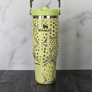 May include: A Stanley brand tumbler with a light green lid and handle. The body of the tumbler is light yellow with a pattern of pickleball graphics. The Stanley logo is visible on the front of the tumbler.