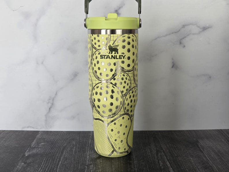 May include: A Stanley brand tumbler with a light green lid and handle. The body of the tumbler is light yellow with a pattern of pickleball graphics. The Stanley logo is visible on the front of the tumbler.