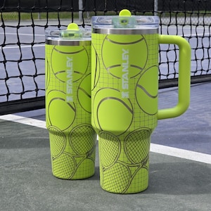 May include: Two lime green Stanley tumblers with tennis ball and racket graphics. One tumbler has a handle. Both have clear lids and green accents. The background includes a tennis net and court.