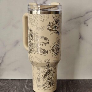 Puede incluir: Un vaso de acero inoxidable beige con un dise&ntilde;o de dinosaurio y flores en blanco y negro. El vaso tiene una tapa y un asa.