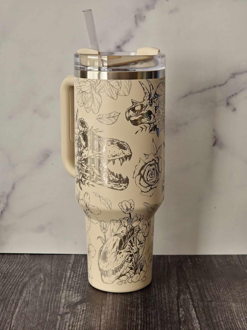Puede incluir: Un vaso de acero inoxidable beige con un dise&ntilde;o de dinosaurio y flores en blanco y negro. El vaso tiene una tapa y un asa.