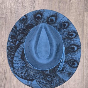 May include: A blue fedora hat with a peacock feather design. The hat has a blue crown and brim, with a black peacock illustration on the brim. A blue tassel hangs from the side.