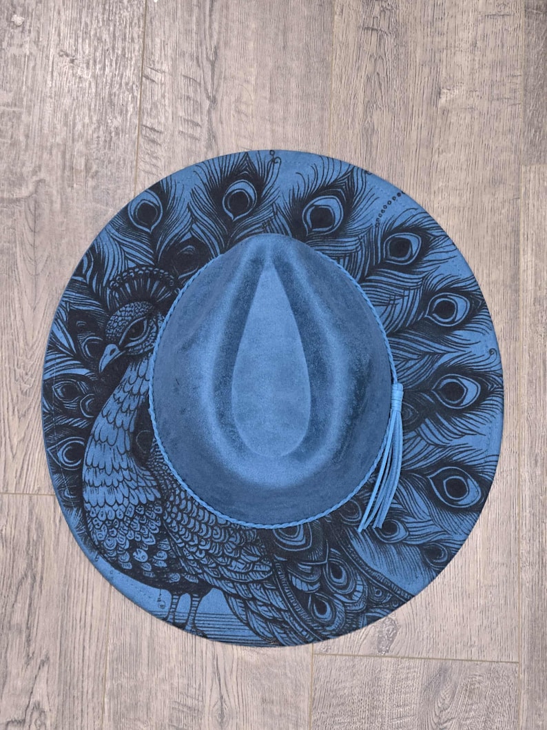 May include: A blue fedora hat with a peacock feather design. The hat has a blue crown and brim, with a black peacock illustration on the brim. A blue tassel hangs from the side.