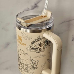 Puede incluir: Una taza de viaje de acero inoxidable de color crema con un dise&ntilde;o floral y calavera en blanco y negro. La taza tiene una tapa con un soporte para pajitas y un asa.