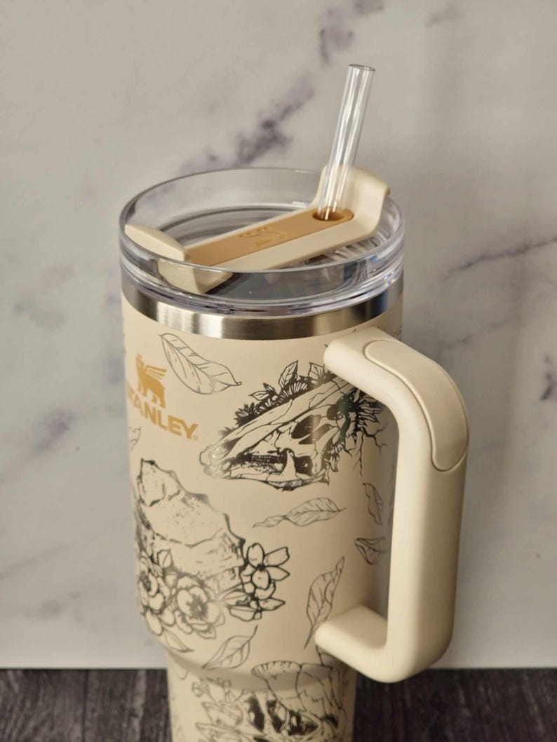 Puede incluir: Una taza de viaje de acero inoxidable de color crema con un dise&ntilde;o floral y calavera en blanco y negro. La taza tiene una tapa con un soporte para pajitas y un asa.