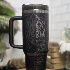 May include: Black travel mug with a handle and a black lid. The mug features the words "ONYX STORM" in a decorative font, along with other text and a map design. The mug is sitting on a wooden surface.