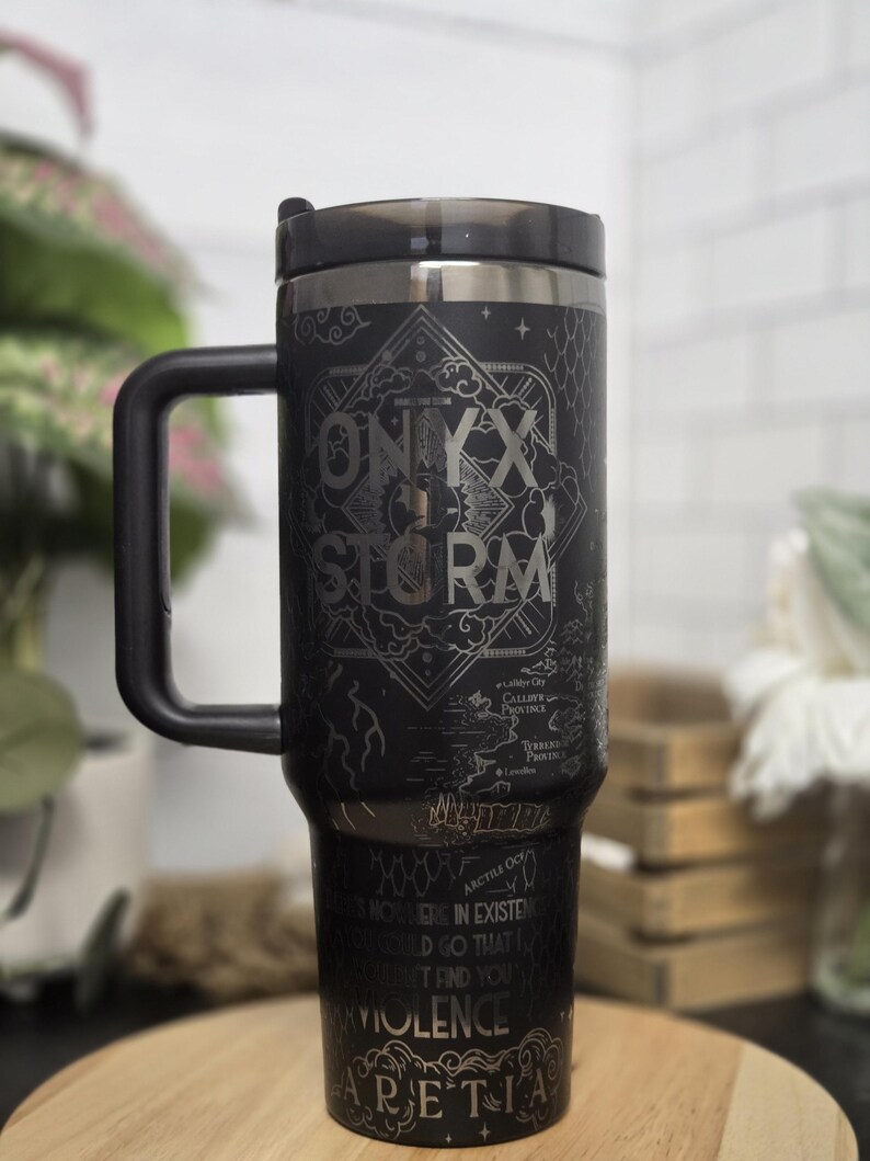 May include: Black travel mug with a handle and a black lid. The mug features the words "ONYX STORM" in a decorative font, along with other text and a map design. The mug is sitting on a wooden surface.