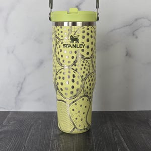 May include: A Stanley tumbler with a light green lid and handle. The body of the tumbler is light yellow with a pattern of silver pickleball-like designs. The Stanley logo is visible on the front.