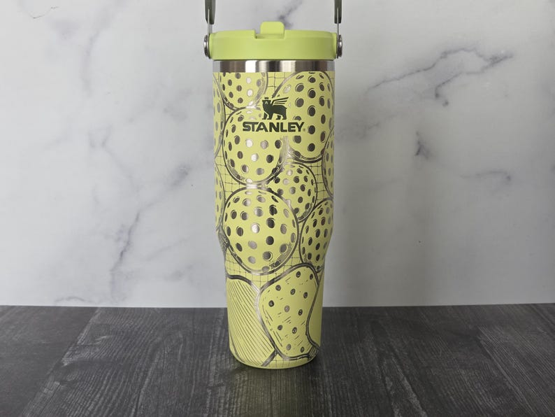 May include: A Stanley tumbler with a light green lid and handle. The body of the tumbler is light yellow with a pattern of silver pickleball-like designs. The Stanley logo is visible on the front.