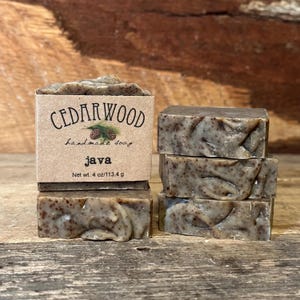 May include: A stack of rectangular bars of speckled brown soap. One bar is wrapped in a tan paper label with the text CEDARWOOD, handmade soap, java, and Net wt. 4 oz/113.4 g. The soap has a textured, marbled appearance with dark brown flecks.