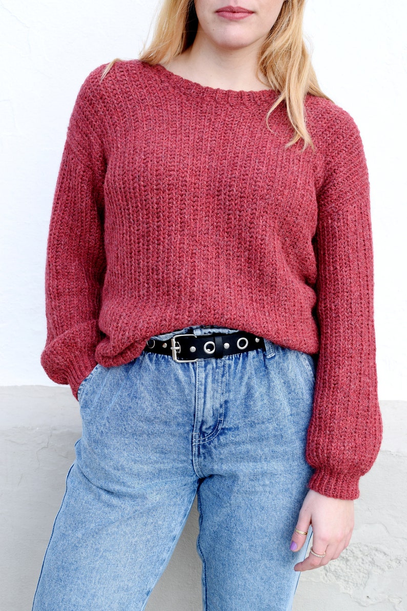 May include: A red knitted sweater with long sleeves and a black belt with silver eyelets. The sweater is worn over a pair of light blue denim jeans.