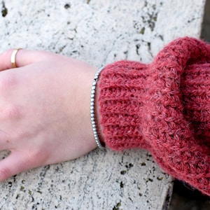 May include: A close-up of a hand wearing a silver beaded bracelet on a wrist. The hand is resting on a textured, light gray surface. The person is wearing two gold rings on their fingers and has purple nail polish.