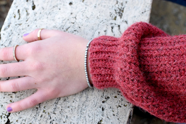 May include: A close-up of a hand wearing a silver beaded bracelet on a wrist. The hand is resting on a textured, light gray surface. The person is wearing two gold rings on their fingers and has purple nail polish.