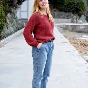 May include: A woman wearing a red knitted sweater, blue jeans, and black shoes. She is standing on a concrete path with a white building and green trees in the background.