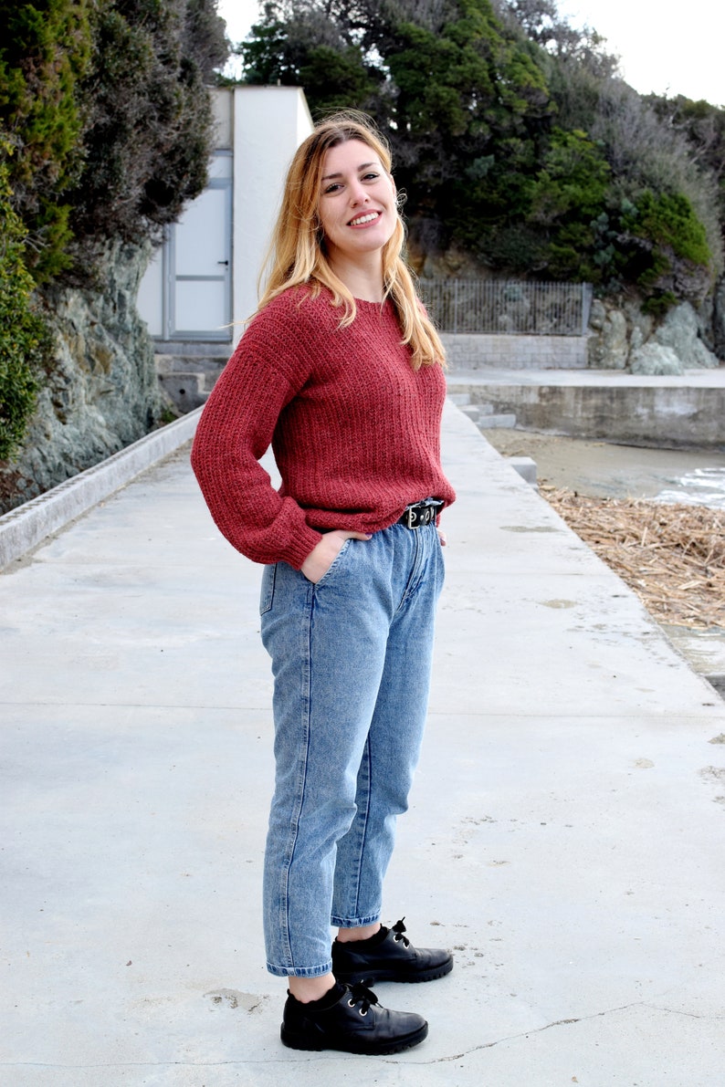 May include: A woman wearing a red knitted sweater, blue jeans, and black shoes. She is standing on a concrete path with a white building and green trees in the background.