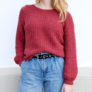 May include: A red knitted sweater with a round neckline and long sleeves. The sweater is worn over a pair of blue jeans with a black belt.