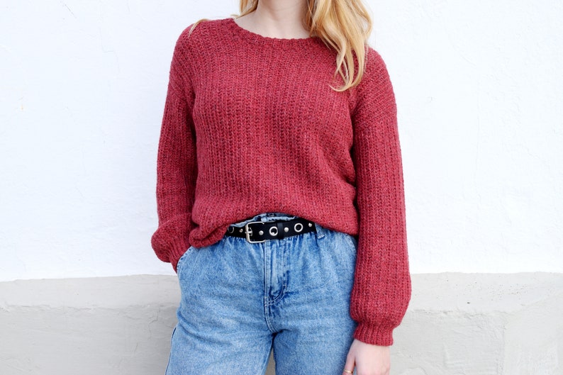 May include: A red knitted sweater with a round neckline and long sleeves. The sweater is worn over a pair of blue jeans with a black belt.