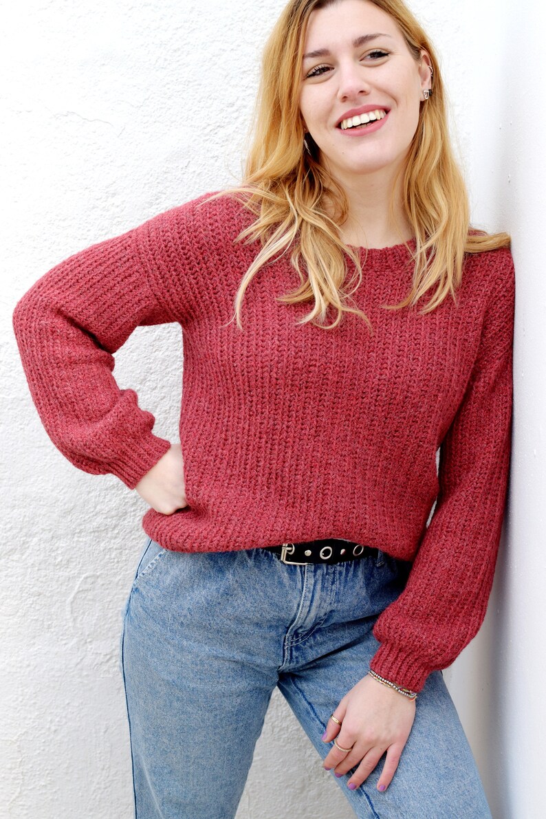May include: A woman wearing a red knitted sweater with a black belt and blue jeans. She is smiling and looking at the camera.