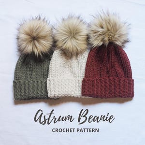 May include: Three crocheted beanies with faux fur pom poms in green, cream, and burgundy. The beanies are arranged in a row on a white surface. The text "Astrum Beanie CROCHET PATTERN" is below the beanies.