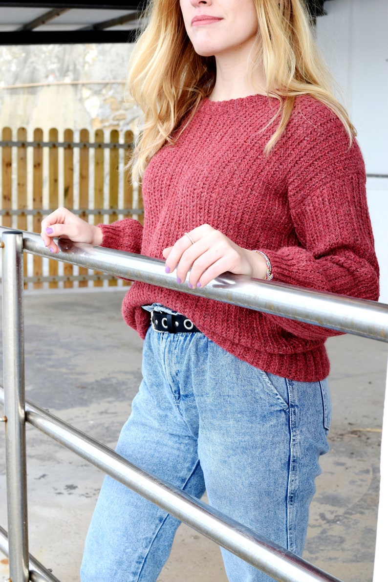 May include: A woman wearing a red knitted sweater with a black belt and blue jeans. She is holding onto a metal railing.