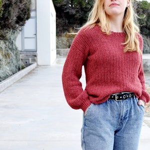 May include: A woman wearing a red knitted sweater with a black belt and blue jeans.