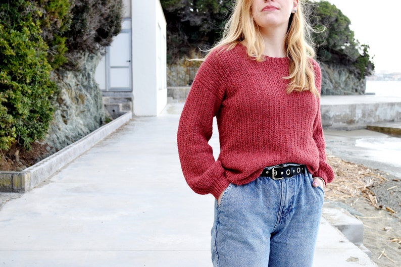 May include: A woman wearing a red knitted sweater with a black belt and blue jeans.