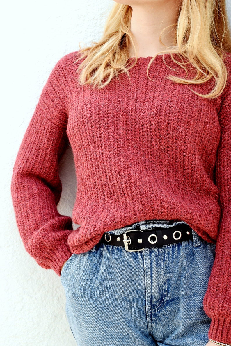 May include: A red knitted sweater with long sleeves and a black belt with silver grommets. The sweater is worn over blue jeans.