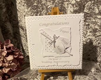 May include: White card with a gray polka dot pattern and the text "Congratulations on your 1st Wedding Anniversary" with a gray scroll and two gray tags with the names "Joseph" and "Katie" on them.